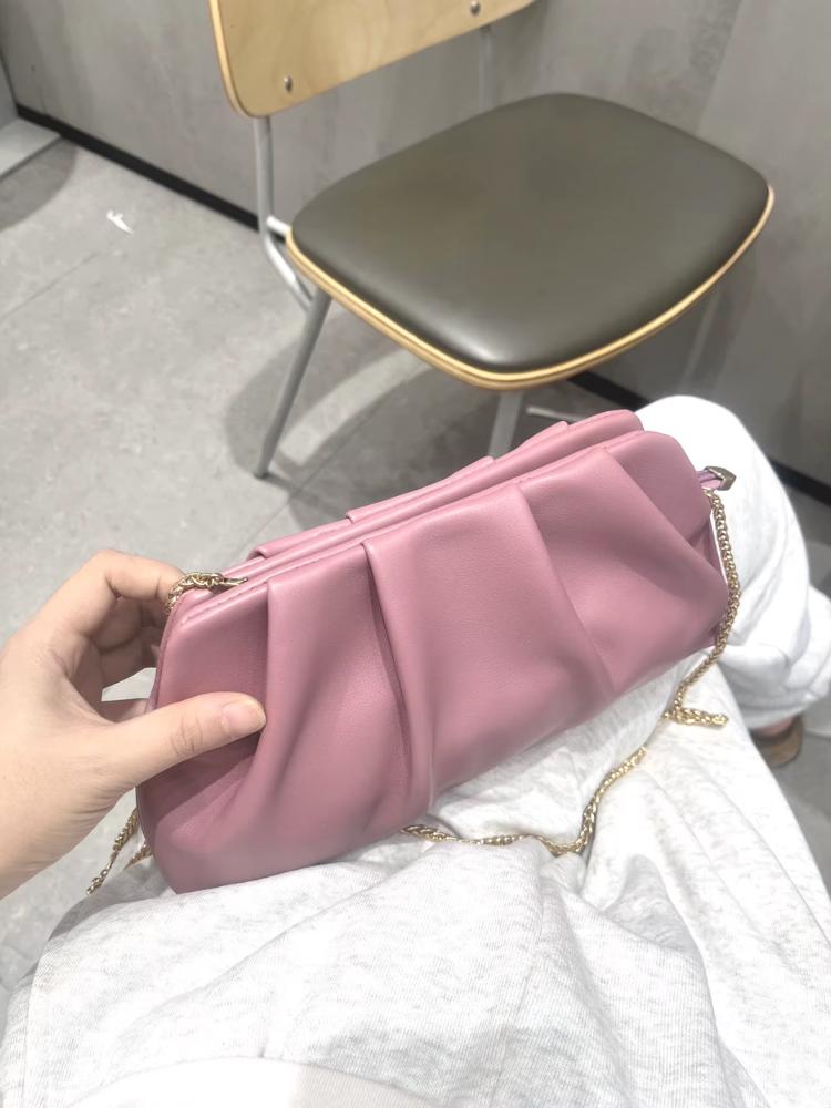 Pessoa segurando bolsa clutch rosa fosca com pregas e corrente dourada no colo, sobre roupa clara.