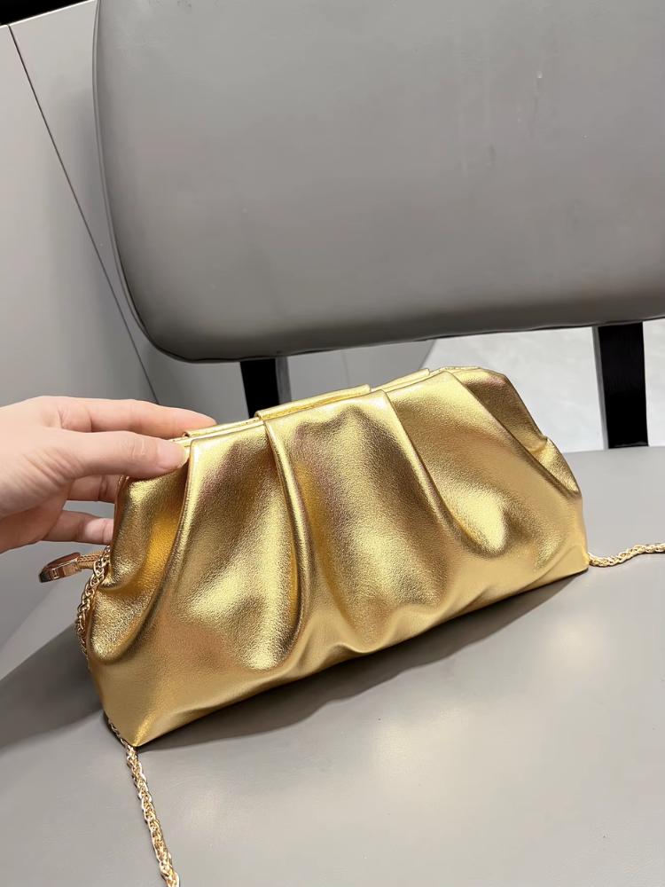 Bolsa clutch dourada com acabamento metalizado, pregas frontais e alça de corrente dourada sobre cadeira cinza.