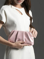 Mulher segurando bolsa clutch rosa metalizada com pregas, combinando com vestido branco elegante.