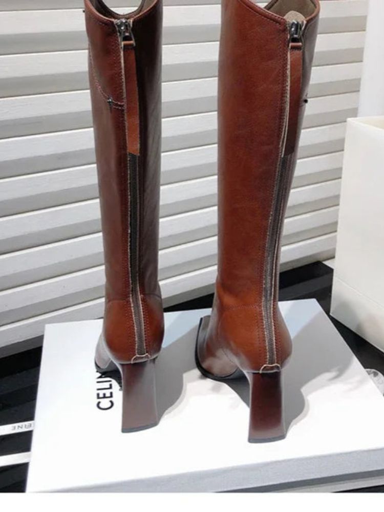 Par de botas femininas de cano alto em couro marrom com bico fino e salto bloco, expostas sobre uma caixa branca.