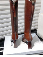 Par de botas femininas de cano alto em couro marrom com bico fino e salto bloco, expostas sobre uma caixa branca.