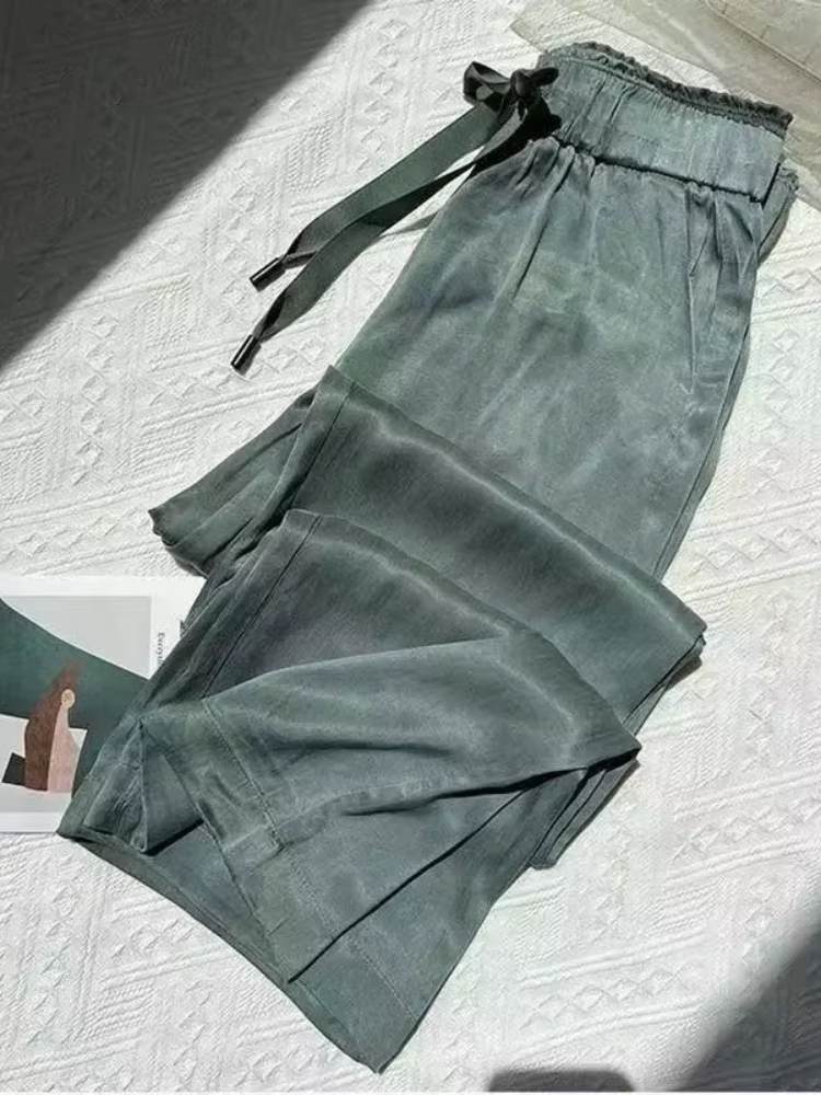Calça feminina ampla em tecido acetinado verde, com cós elástico e laço frontal, dobrada sobre superfície clara.