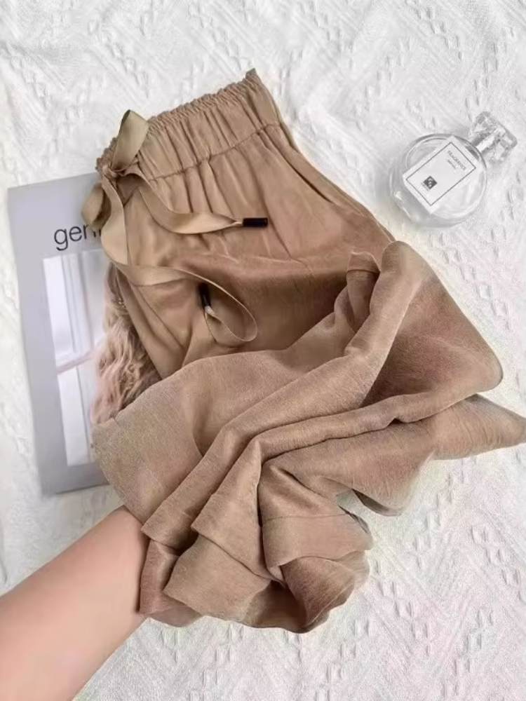 Calça feminina bege em tecido leve e acetinado, com cós elástico e laço, sendo segurada para mostrar o caimento fluido.