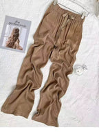 Calça feminina bege ampla esticada sobre superfície branca, mostrando caimento leve e tecido brilhante.