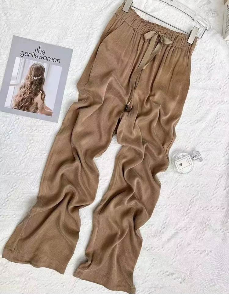 Calça feminina bege ampla esticada sobre superfície branca, mostrando caimento leve e tecido brilhante.