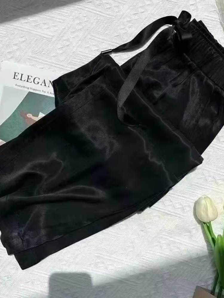 Calça feminina preta em tecido acetinado, dobrada sobre superfície clara com detalhe de laço na cintura.