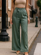 Modelo usando calça verde ampla de tecido acetinado, cintura alta com amarração frontal, em ambiente urbano.
