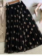 Saia longa de tule preta com corações bege estendida ao lado de blazer e sapatos nude.