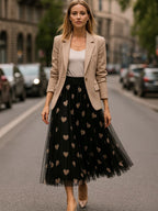 Saia longa de tule preta com estampa de corações bege, combinada com blazer bege e blusa branca em rua da cidade.