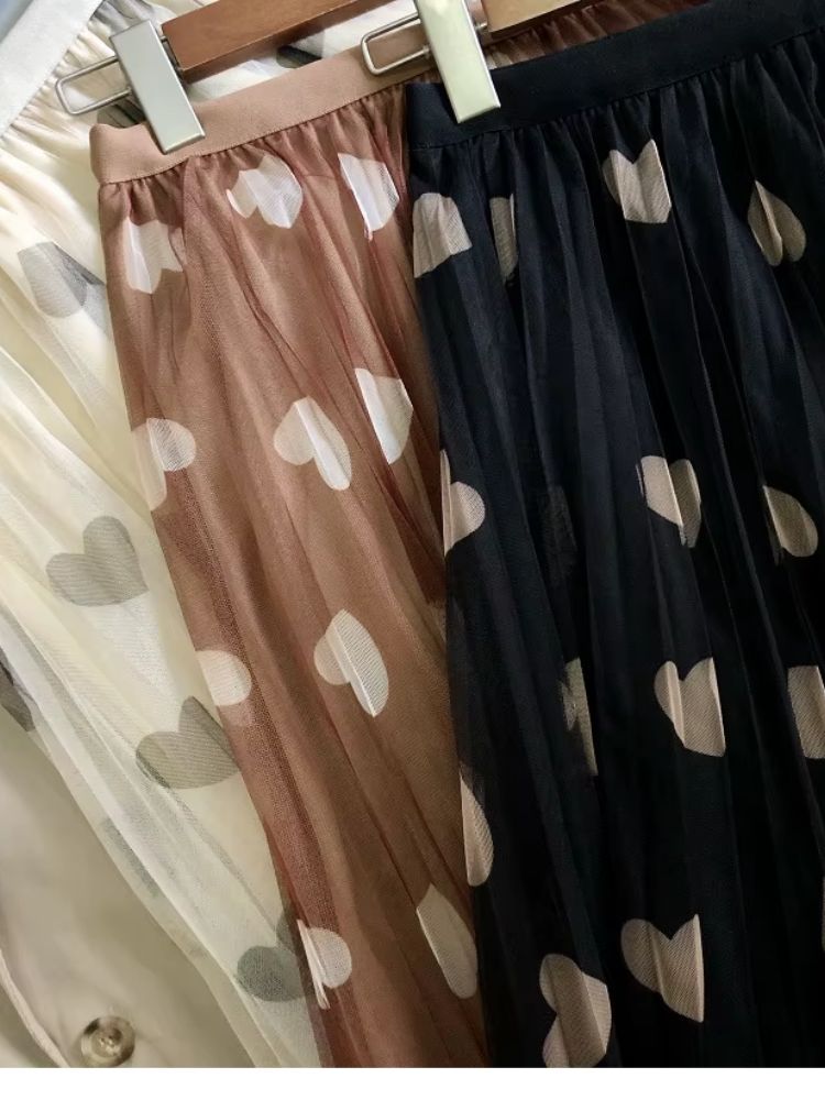 Três saias longas de tule com estampa de corações — bege, marrom e preto — penduradas lado a lado em cabides.