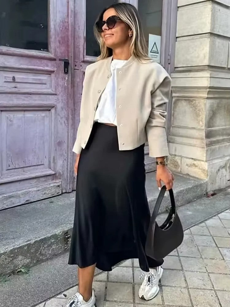 Saia midi preta em cetim com caimento fluido, usada com jaqueta clara, tênis esportivo e bolsa estruturada preta em look urbano moderno.