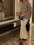 Saia midi branca em cetim com brilho suave, combinada com suéter bege oversized e sandálias pretas em ambiente sofisticado.