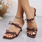 Sandália vinho de salto médio com tiras finas e laços frontais, modelo aberto no calcanhar, exibida nos pés com tornozeleira decorativa.