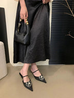 Sandálias femininas pretas de bico fino com aplique de flor e pérola, usadas com saia midi preta texturizada e bolsa preta pequena de alça com esferas douradas